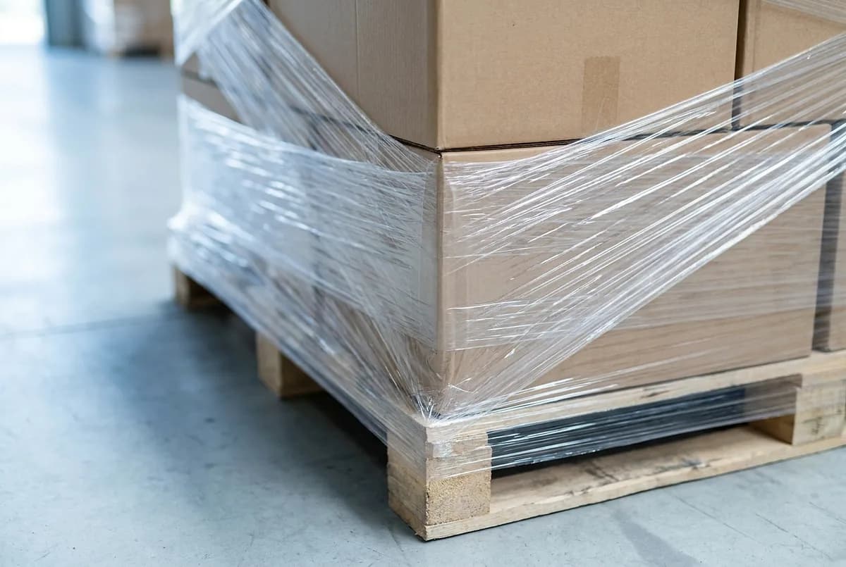 The corner of a carton pallet wrapped in several layers of clear stretch film, with the tension lines visible along the edge and under the pallet